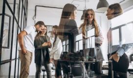 A group of six people stand and talk in a modern office, some discussing software development methodologies while others use phones near large windows and glass walls.