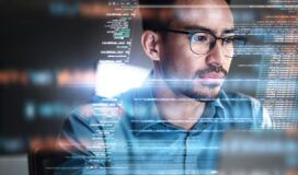 A man wearing glasses looks at a computer screen displaying lines of code, suggesting he is working on programming or developing SaaS applications.