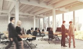 Modern open-plan office with large windows, empty desks with computers, two people discussing enterprise application development, and two others walking past in motion blur.