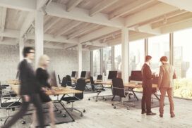 Modern open-plan office with large windows, empty desks with computers, two people discussing enterprise application development, and two others walking past in motion blur.
