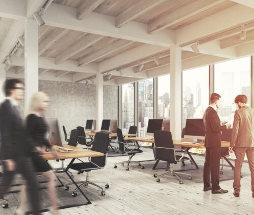 Modern open-plan office with large windows, empty desks with computers, two people discussing enterprise application development, and two others walking past in motion blur.