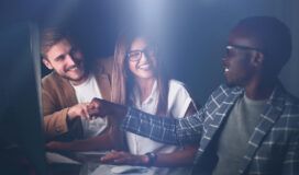 Three people in business attire work at computers, smiling and giving a fist bump, celebrating success with their software development partner in an office setting with soft lighting.