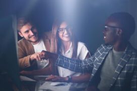 Three people in business attire work at computers, smiling and giving a fist bump, celebrating success with their software development partner in an office setting with soft lighting.