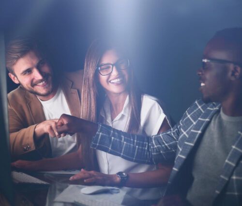Three people in business attire work at computers, smiling and giving a fist bump, celebrating success with their software development partner in an office setting with soft lighting.