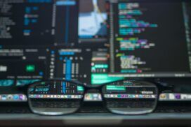 A pair of eyeglasses sits on a laptop keyboard, with code and digital interfaces visible on the laptop screen in the background.