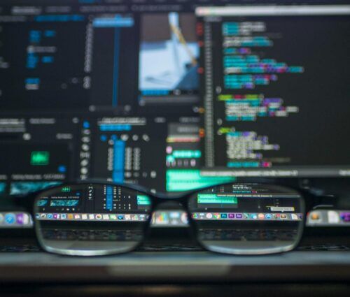 A pair of eyeglasses sits on a laptop keyboard, with code and digital interfaces visible on the laptop screen in the background.
