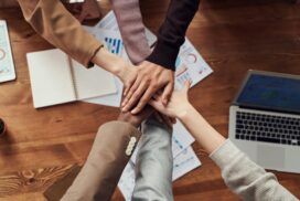 Five people stack their hands together over a wooden table with documents, a notebook, and a laptop, symbolizing teamwork and the strong collaboration needed when choosing a software development partner.