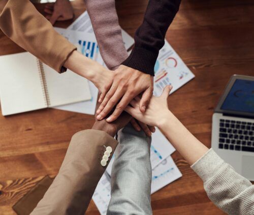 Five people stack their hands together over a wooden table with documents, a notebook, and a laptop, symbolizing teamwork and the strong collaboration needed when choosing a software development partner.