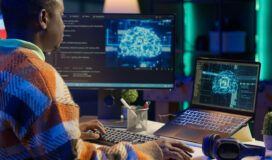 A person works at a desk with a desktop monitor and laptop displaying digital illustrations of a brain, surrounded by code and technological elements, highlighting Palantir consulting and implementation services in action.