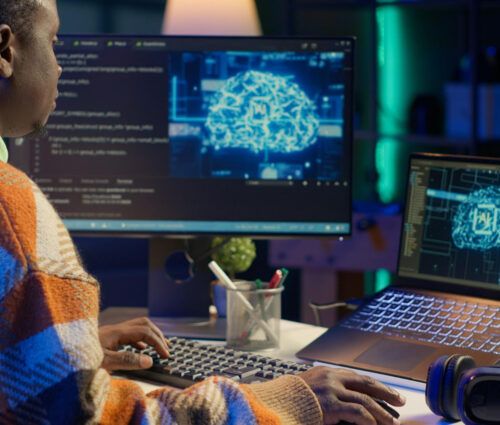 A person works at a desk with a desktop monitor and laptop displaying digital illustrations of a brain, surrounded by code and technological elements, highlighting Palantir consulting and implementation services in action.