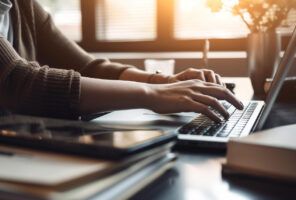 Person typing on a laptop at a desk with books, a tablet, and a pen in a sunlit room.