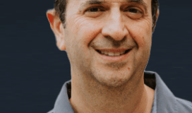 Middle-aged man with short dark hair and a gray collared shirt, smiling in front of a plain dark background.