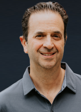 Middle-aged man with short dark hair and a gray collared shirt, smiling in front of a plain dark background.