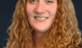 A woman with long, curly red hair, wearing a black blazer and light-colored top, poses in front of a dark background, smiling at the camera.