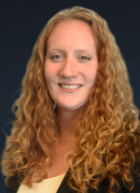 A woman with long, curly red hair, wearing a black blazer and light-colored top, poses in front of a dark background, smiling at the camera.