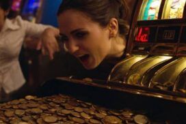 A woman leans close to a slot machine overflowing with coins, appearing excited while another person reacts enthusiastically in the background.