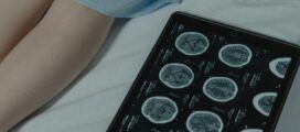 A tablet displaying brain scan images lies on a bed next to a person's legs, partially covered by a hospital gown.