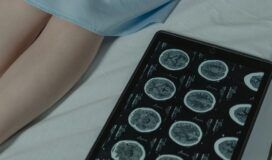 A tablet displaying brain scan images lies on a bed next to a person's legs, partially covered by a hospital gown.