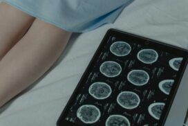 A tablet displaying brain scan images lies on a bed next to a person's legs, partially covered by a hospital gown.