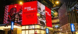 A busy city street at night with people walking and cars passing in front of a brightly lit shopping mall displaying a large red "Icons Craft" advertisement.