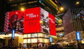 A busy city street at night with people walking and cars passing in front of a brightly lit shopping mall displaying a large red "Icons Craft" advertisement.