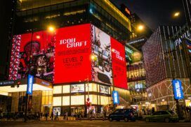 A busy city street at night with people walking and cars passing in front of a brightly lit shopping mall displaying a large red "Icons Craft" advertisement.