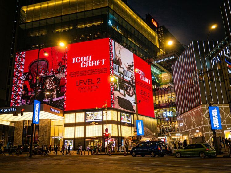 A busy city street at night with people walking and cars passing in front of a brightly lit shopping mall displaying a large red "Icons Craft" advertisement.