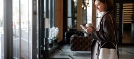 Woman in a brown coat holding a coffee cup and looking at her phone stands near the entrance of a modern building with glass doors and contemporary decor.