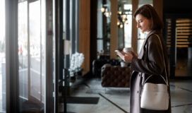 Woman in a brown coat holding a coffee cup and looking at her phone stands near the entrance of a modern building with glass doors and contemporary decor.