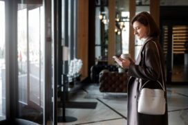 Woman in a brown coat holding a coffee cup and looking at her phone stands near the entrance of a modern building with glass doors and contemporary decor.
