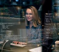 A woman wearing glasses works at a computer with multiple lines of code and an AI coding assistant displayed on the screen. A takeout coffee cup is on the desk beside her.