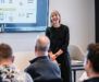 A woman stands and smiles while presenting data on a screen to an audience seated in a modern conference room.