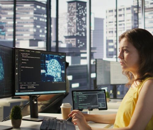A woman in a yellow top works at a desk with multiple monitors displaying code and a digital brain image, illustrating the development of ai agent services in a modern office with large windows and city views.