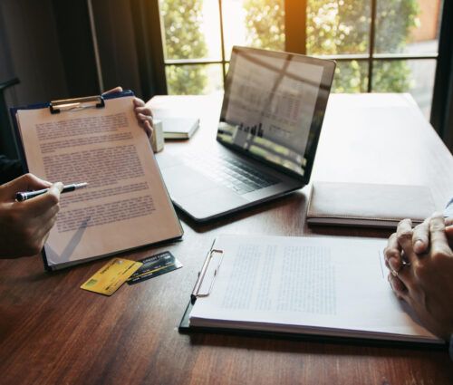 Two people sit at a wooden table with documents, credit cards, a laptop, and a notebook during a meeting to discuss ai agent services.