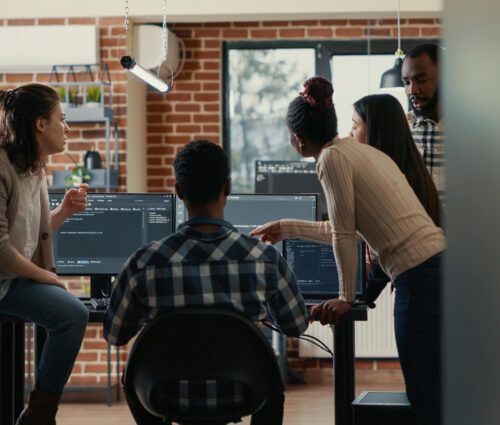 Four people collaborate around computer monitors displaying code and discussing ai agent services in a modern office with brick walls and large windows.