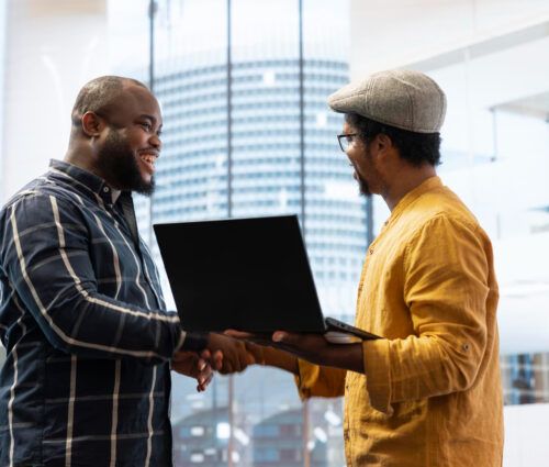 Two men stand indoors, one holding a laptop, as they smile and shake hands in a modern office setting—highlighting a successful collaboration on AI agent services with city buildings visible through large windows.