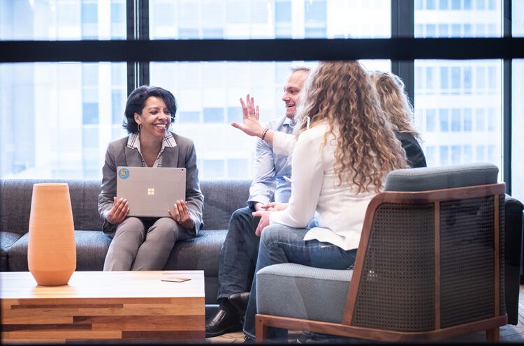 Four people sit on couches in a modern office, engaged in conversation. One person holds a laptop while another gestures enthusiastically. A table and vase are in the foreground.