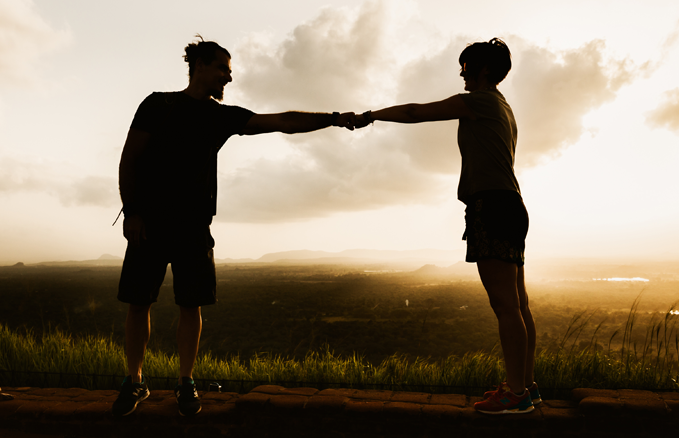 Silhouettes of two people standing outdoors at sunset, facing each other and touching fists, with a scenic landscape in the background.