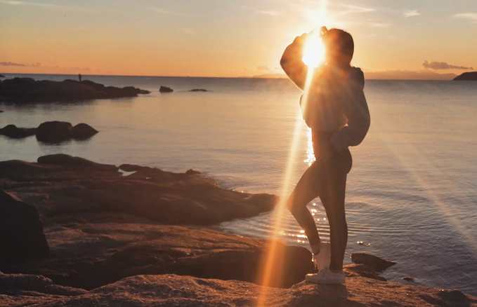 Person standing on rocky shore at sunset with the sun shining behind them, casting a lens flare across the image; calm water and rocks in the background.