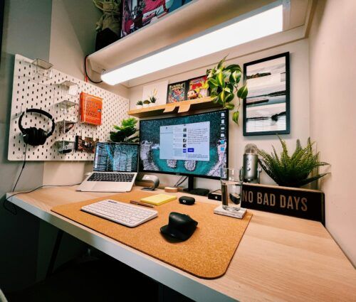 A neatly organized home office desk with a laptop, monitor, keyboard, mouse, plants, headphones, and a sign that reads "NO BAD DAYS.