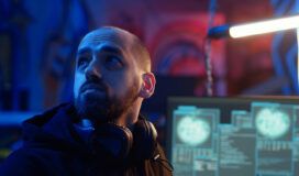 A man with a beard and headphones around his neck looks to the side in a dimly lit room with computer monitors displaying data in the background.