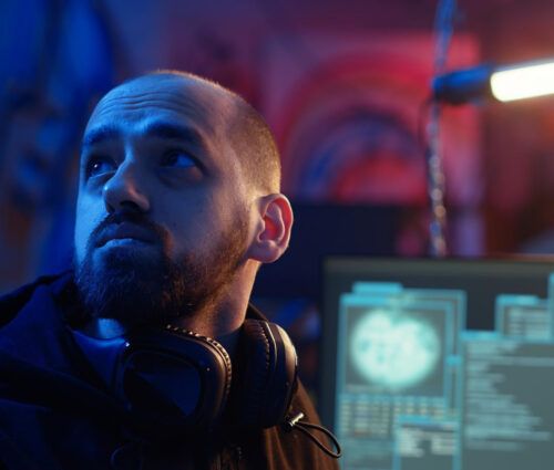 A man with a beard and headphones around his neck looks to the side in a dimly lit room with computer monitors displaying data in the background.