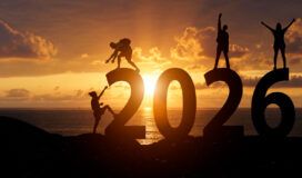 Four people pose on large "2026" numerals at sunset by the sea, silhouetted against an orange sky—capturing a moment of anticipation for 2025 tech predictions as the sun meets the horizon.