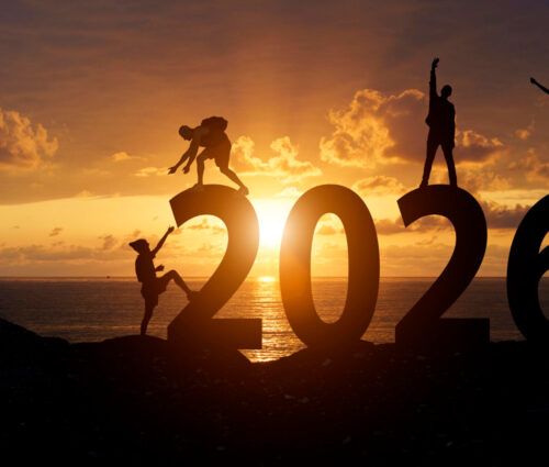 Four people pose on large "2026" numerals at sunset by the sea, silhouetted against an orange sky—capturing a moment of anticipation for 2025 tech predictions as the sun meets the horizon.