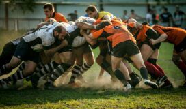 Two rugby teams, one in white and the other in orange, engage in a scrum on a grassy field during a match, moments before a potential scrum failure.