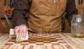 A person in a brown apron wipes oil onto a wooden chessboard in a workshop, with tools and AI tools on the wall, and a jar of oil on the table.