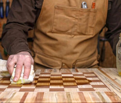 A person in a brown apron wipes oil onto a wooden chessboard in a workshop, with tools and AI tools on the wall, and a jar of oil on the table.