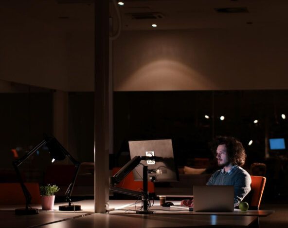 A man sits alone at a desk in a dimly lit office at night, working on a laptop with a plant and an apple nearby—researching AI for business leaders.
