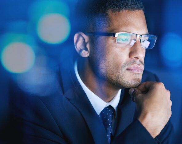 A man in a suit and glasses looks intently at a computer screen, resting his chin on his hand, exploring Ai for business leaders against a blurred blue background.