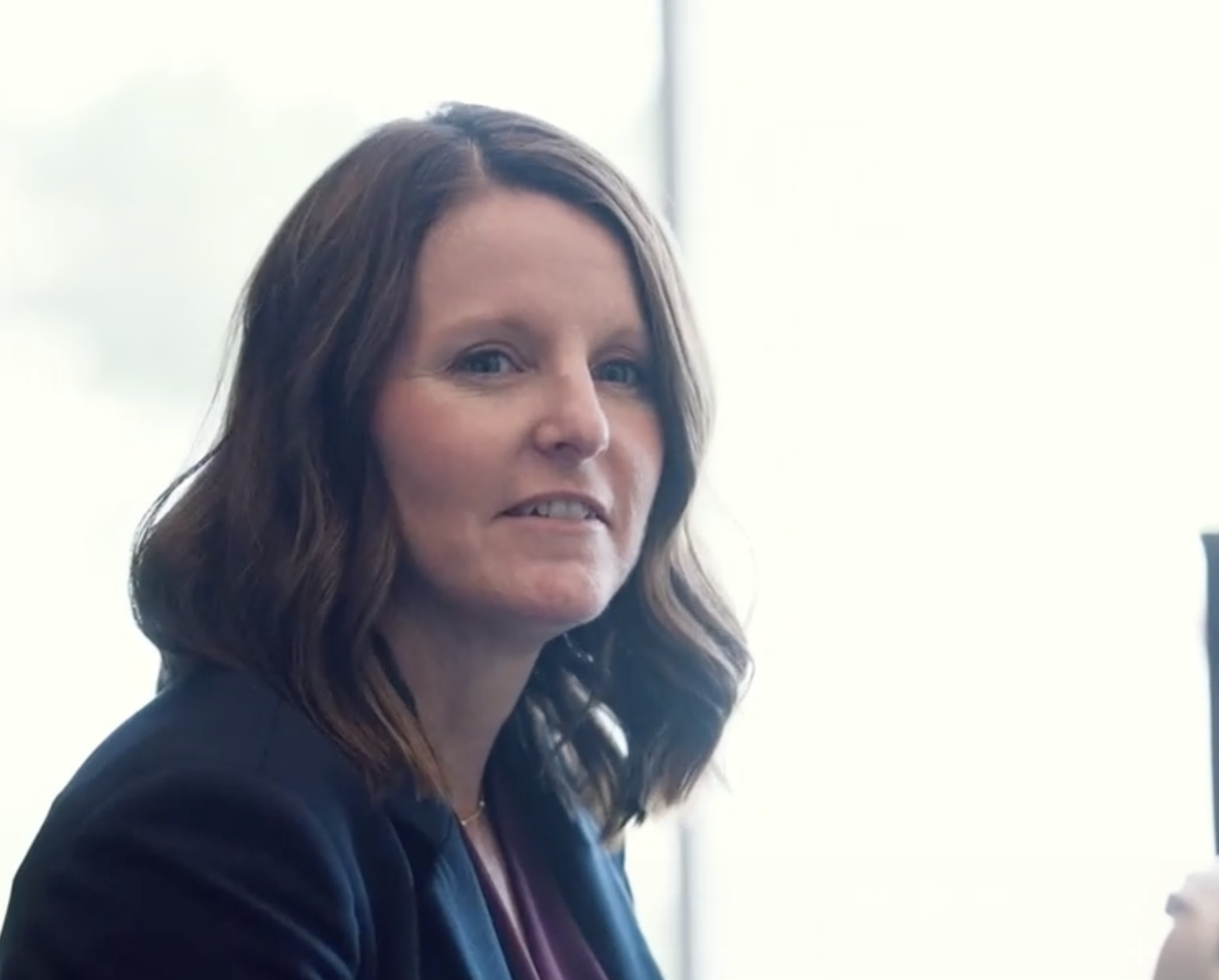 A woman with shoulder-length brown hair and a navy blazer sits indoors, looking toward the camera with a neutral expression, suggesting confidence in her expertise in ML for insurance.